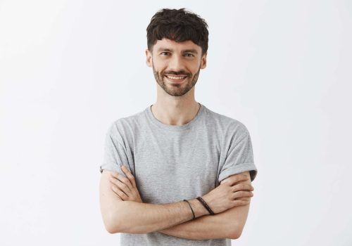 Waist-up shot of confident good-looking mature european male entrepreneur in t-shirt holding hands crossed in self-assured pose and smiling with assured pleasant smile over gray background.