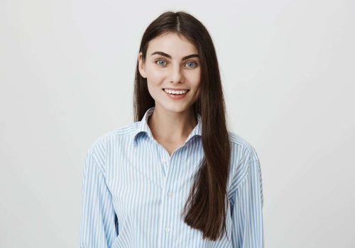 Waist-up portrait of happy and confident brunette woman smiling broadly at camera while standing over gray background. Office worker greets customers and offers some tea or coffee. Emotions concept.